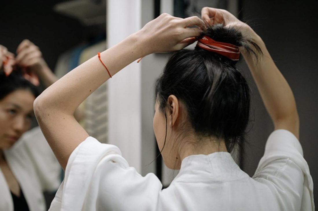 A woman ties her hair in a bun in front of the mirror. (From: Pexels)