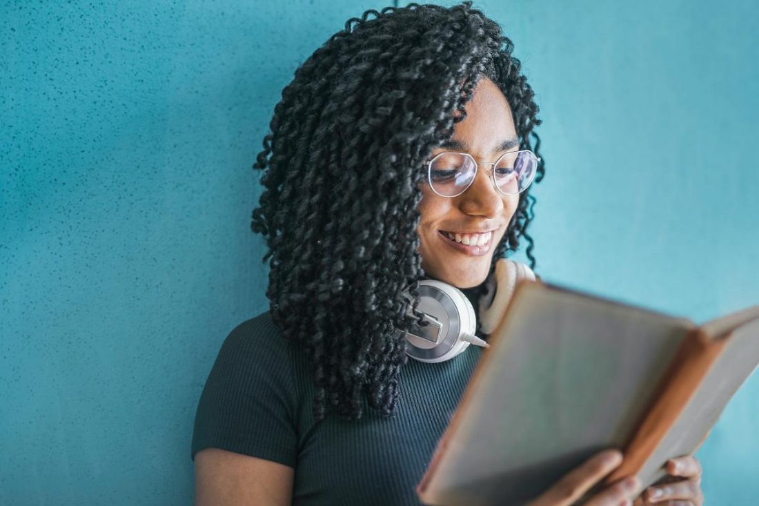 Woman reading a book with headphones around her neck. (From: Pexels)