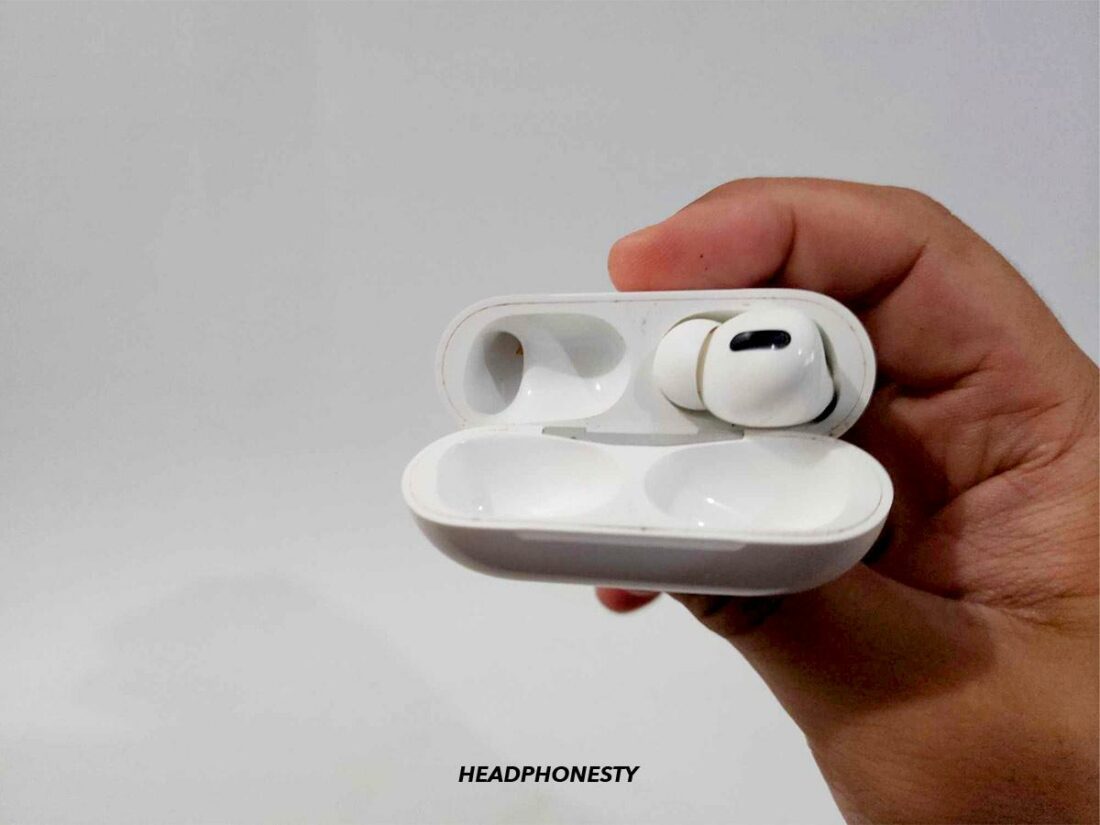 The left AirPod inside the charging case with the lid open while pressing the setup button to reset.